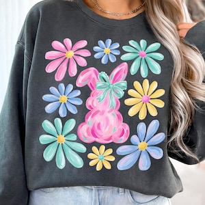 May include: Dark gray sweatshirt featuring a pink bunny with a teal bow, surrounded by colorful flowers. The flowers are in shades of pink, blue, yellow, and teal. A casual, springtime themed top.