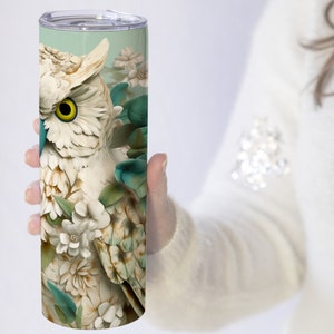 May include: A white ceramic owl with teal and green floral accents on a stainless steel tumbler. The owl has a yellow eye and is surrounded by white flowers.