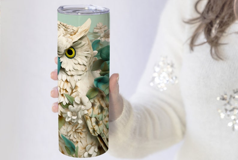 May include: A white ceramic owl with teal and green floral accents on a stainless steel tumbler. The owl has a yellow eye and is surrounded by white flowers.