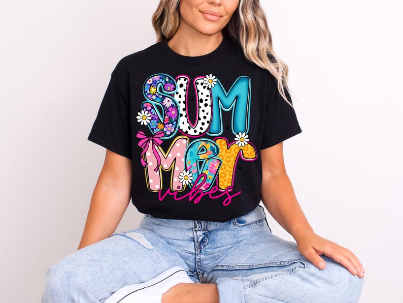 May include: Black t-shirt with the words "SUMMER VIBES" in a colorful, playful design. The letters are decorated with floral patterns, polka dots, and animal print, with daisy accents. A pink bow is also included.