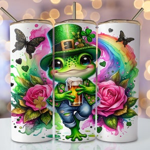 Puede incluir: Un vaso blanco con una rana verde que lleva un sombrero de duende verde y sostiene un vaso de cerveza. El fondo presenta un arcoíris, rosas rosas y tréboles verdes.
