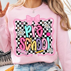 May include: Pink sweatshirt with a colorful "Pre School" graphic. The design features a pink bow, daisies, a checkered background, and a pencil. The text is in various colors, including blue, orange, and purple. A cute and fun design for preschool.