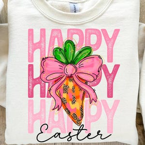 Puede incluir: Una sudadera blanca con las palabras "Happy Happy Happy Easter" en letras rosas. Una zanahoria de dibujos animados con un lazo rosa y hojas verdes está en el centro del texto.