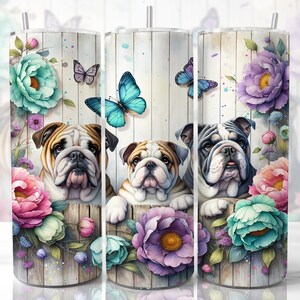 Op de afbeelding: Drie Engelse Bulldogs die uit een bloemstuk kijken. De bloemen zijn roze, paars en blauw met een houten achtergrond. De honden zijn wit met zwarte vlekken en hebben een lieve uitdrukking.
