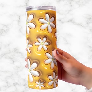 May include: A tall, cylindrical tumbler with a clear lid. The tumbler features a repeating pattern of white daisies with yellow centers against a glossy, golden-yellow background. The tumbler is held by a person's hand.