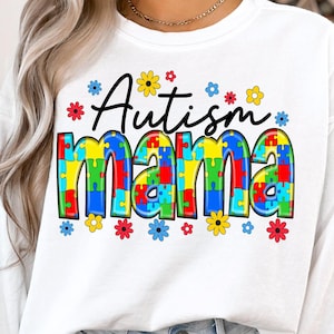 Puede incluir: Camiseta blanca de manga larga con el texto "Autism Mama" en letras negras. La palabra "Mama" está formada por piezas de rompecabezas de colores y rodeada de pequeñas flores amarillas, rojas, azules y blancas.