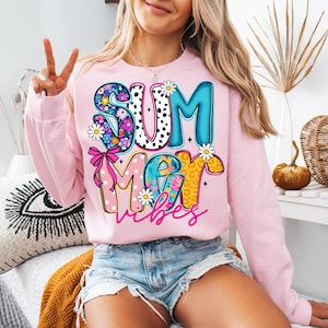 May include: A light pink sweatshirt with the words "SUMMER vibes" in a colorful, playful design. The letters are decorated with floral, polka dot, and leopard print patterns. Daisies and a pink bow accent the design.