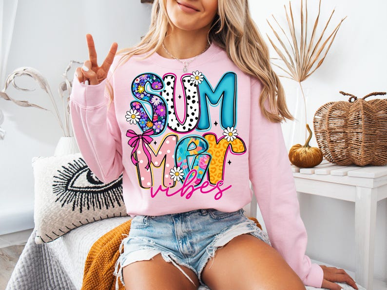 May include: A light pink sweatshirt with the words "SUMMER vibes" in a colorful, playful design. The letters are decorated with floral, polka dot, and leopard print patterns. Daisies and a pink bow accent the design.