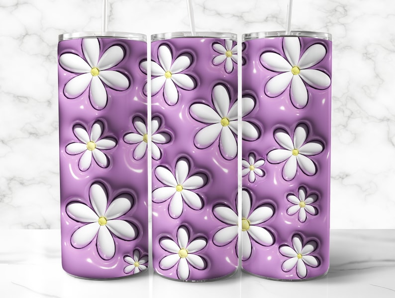 May include: A set of three stainless steel tumblers with a floral design. The design features white daisies with yellow centers against a glossy, lavender background. The tumblers have a straw.