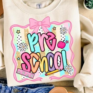 May include: Cream-colored sweatshirt with a colorful "Pre School" graphic. The design features a pink bow, stars, flowers, and a ruler. The text is in various colors, including teal, pink, orange, and purple. A cute and colorful design for preschool.