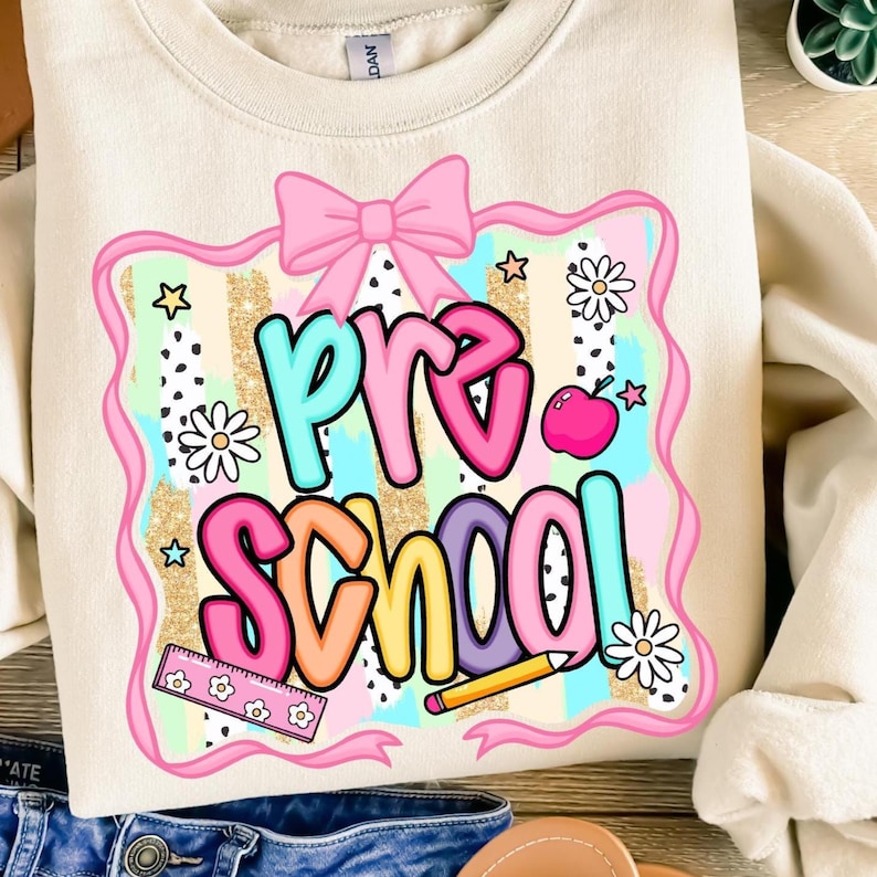 May include: Cream-colored sweatshirt with a colorful "Pre School" graphic. The design features a pink bow, stars, flowers, and a ruler. The text is in various colors, including teal, pink, orange, and purple. A cute and colorful design for preschool.