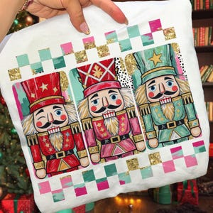 May include: White decorative pillow featuring three colorful nutcracker soldiers. Each nutcracker has a unique uniform in red, pink, and green, with gold accents and a star on their hats. The pillow has a checkered border.