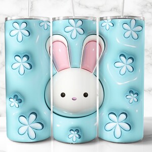 May include: A light blue tumbler with a white bunny face and pink ears. The tumbler has a 3D floral pattern and a clear straw. The design is repeated on the tumbler's surface.