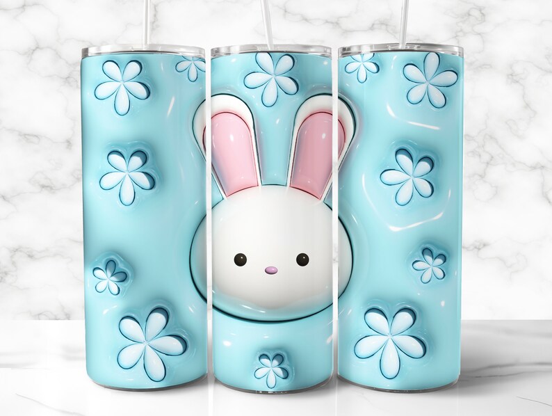 May include: A light blue tumbler with a white bunny face and pink ears. The tumbler has a 3D floral pattern and a clear straw. The design is repeated on the tumbler's surface.