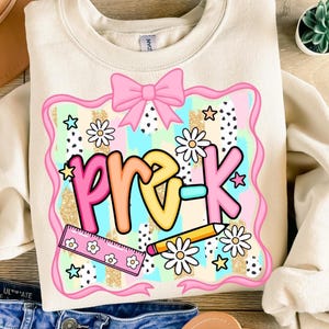 May include: Cream-colored sweatshirt with a colorful "pre-K" graphic. The graphic includes a pink bow, daisies, stars, a ruler, and a pencil. The letters are in pink, yellow, and blue. The design is framed with a pink border.