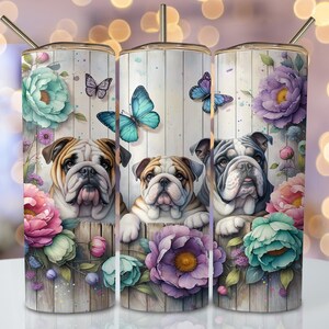 Op de afbeelding: Drie roestvrijstalen bekers met een bloemenontwerp en afbeeldingen van Engelse Bulldogs. De bekers hebben een witte houtnerf achtergrond met roze, paarse en blauwe bloemen. De Bulldogs kijken door de bloemen.