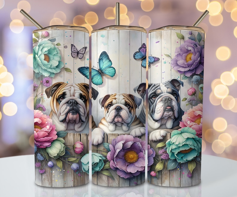 Op de afbeelding: Drie roestvrijstalen bekers met een bloemenontwerp en afbeeldingen van Engelse Bulldogs. De bekers hebben een witte houtnerf achtergrond met roze, paarse en blauwe bloemen. De Bulldogs kijken door de bloemen.