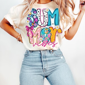 May include: Cream-colored t-shirt with the words "SUMMER VIBES" in a colorful, floral, and patterned design. The word "SUM" is in blue and black polka dots, "MER" is pink, and "VIBES" is in a cursive font.