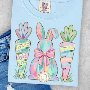May include: Light blue t-shirt featuring a pastel bunny with a pink bow and two carrots. The bunny and carrots have a colorful, leopard-print design. The shirt has the Comfort Colors brand tag.