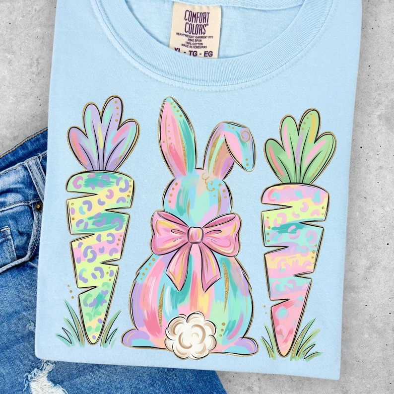 May include: Light blue t-shirt featuring a pastel bunny with a pink bow and two carrots. The bunny and carrots have a colorful, leopard-print design. The shirt has the Comfort Colors brand tag.