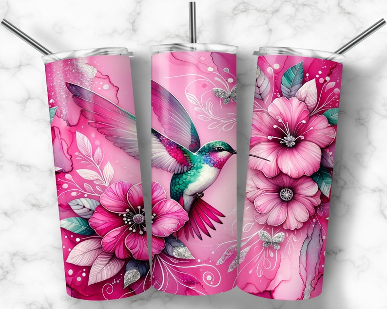 Pode incluir: Um copo rosa com um design vibrante de beija-flor. O copo apresenta um estilo aquarela com flores rosa, folhas e borboletas. O beija-flor tem penas coloridas. Inclui um canudo prateado.