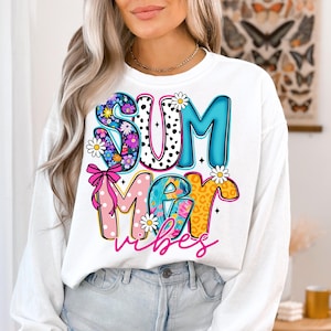 May include: White sweatshirt with the words "SUMMER VIBES" in a colorful, playful design. The letters are decorated with floral patterns, polka dots, and leopard print. A pink bow and daisies add to the summery theme.