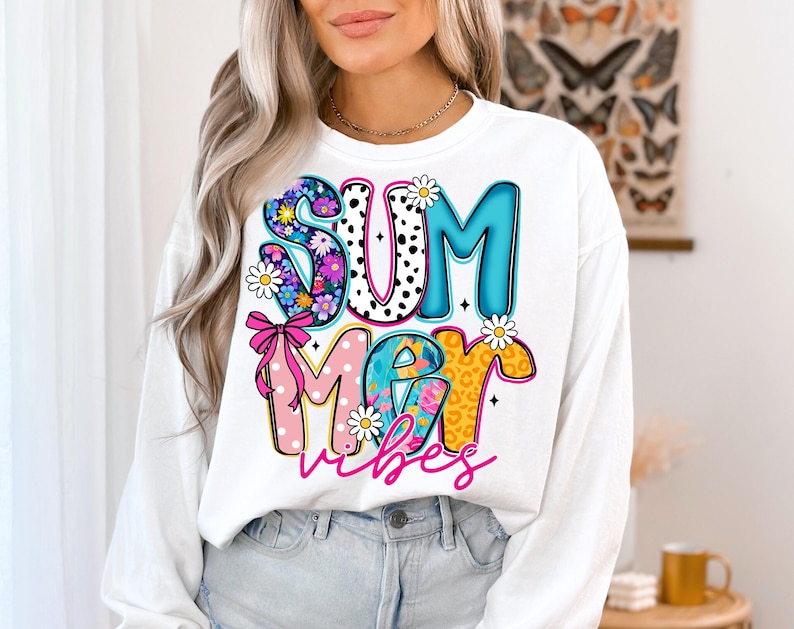 May include: White sweatshirt with the words "SUMMER VIBES" in a colorful, playful design. The letters are decorated with floral patterns, polka dots, and leopard print. A pink bow and daisies add to the summery theme.