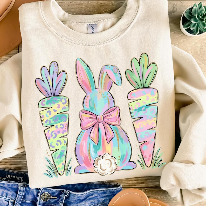 May include: Cream-colored sweatshirt featuring a pastel bunny with a pink bow and two carrots. The bunny and carrots have a colorful, leopard-print design. The design is set against a cream background, with green grass at the bottom.