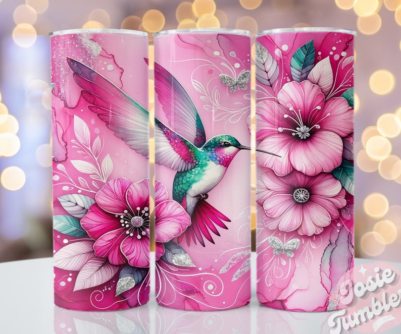 Pode incluir: Um copo rosa com um desenho de um beija-flor com penas rosa, turquesa e roxas, junto a grandes flores rosas e detalhes de glitter prateado. O design inclui elementos florais e de folhas, com o texto "Josie Tumbler".