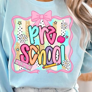 May include: Light blue sweatshirt with a colorful "Pre School" graphic. The design includes a pink bow, stars, daisies, a ruler, a pencil, and an apple. The text is in various colors, outlined in black.