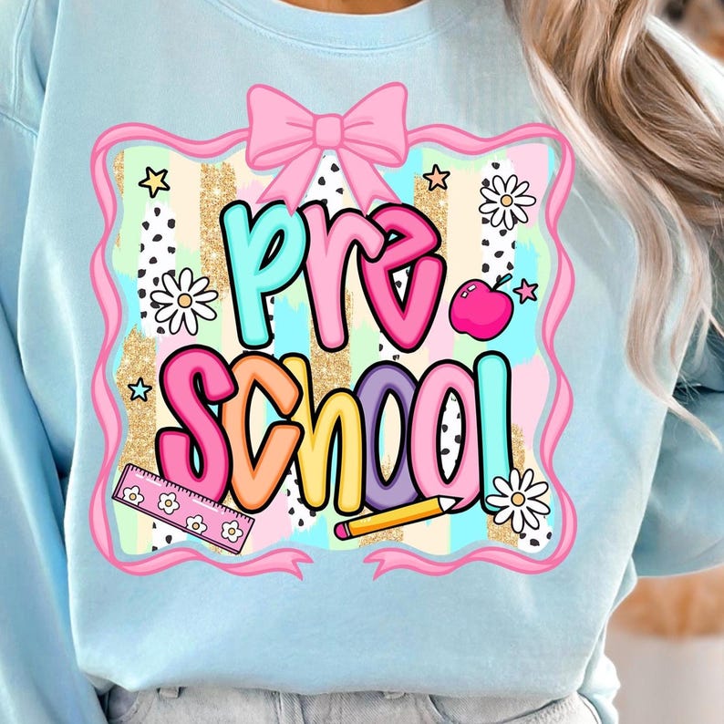 May include: Light blue sweatshirt with a colorful "Pre School" graphic. The design includes a pink bow, stars, daisies, a ruler, a pencil, and an apple. The text is in various colors, outlined in black.