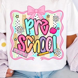 May include: White t-shirt featuring a colorful "Pre School" design. The design includes a pink bow, stars, daisies, a ruler, a pencil, and an apple. The text is in various colors, with a glittery gold background.