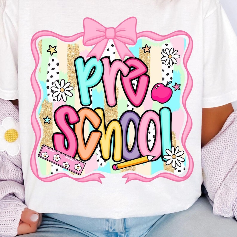 May include: White t-shirt featuring a colorful "Pre School" design. The design includes a pink bow, stars, daisies, a ruler, a pencil, and an apple. The text is in various colors, with a glittery gold background.