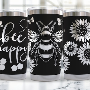May include: Three stainless steel tumblers with black backgrounds and silver designs. One tumbler features the text "bee happy" with honeycomb designs, another has a bee illustration, and the third has a sunflower design.