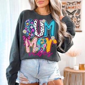 May include: Dark gray sweatshirt with a colorful "SUMMER VIBES" graphic. The word "SUMMER" is in a mix of floral, polka dot, and leopard print patterns. The word "VIBES" is in a cursive font. White daisy accents.