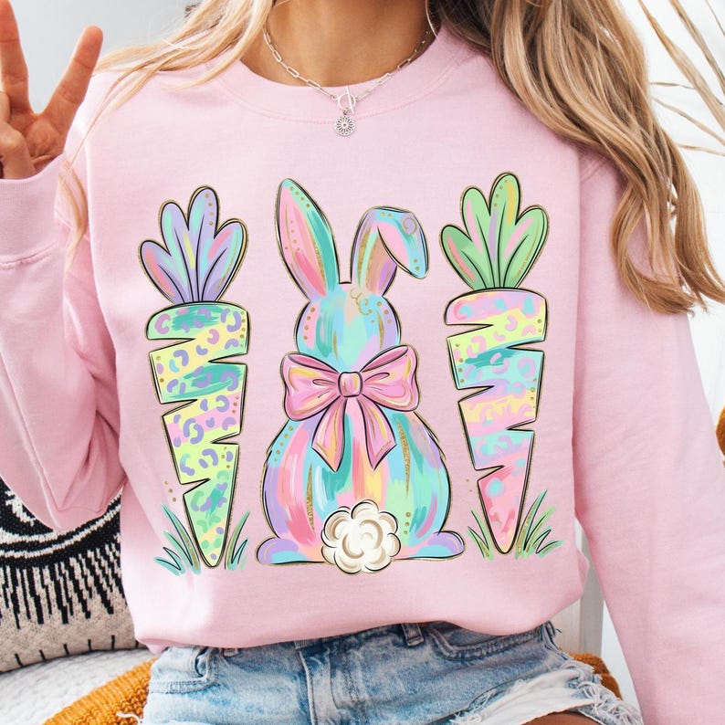May include: A light pink sweatshirt featuring a pastel bunny with a bow and two carrots. The bunny and carrots have a colorful design. The design is set against a light pink background, creating a cheerful and festive look.