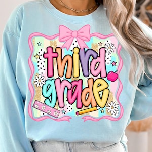 May include: Light blue sweatshirt with a colorful "third grade" graphic. The design includes a pink bow, flowers, a ruler, a pencil, and an apple. The text is in a playful, multi-colored font, perfect for back-to-school.