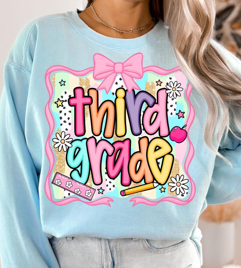 May include: Light blue sweatshirt with a colorful "third grade" graphic. The design includes a pink bow, flowers, a ruler, a pencil, and an apple. The text is in a playful, multi-colored font, perfect for back-to-school.