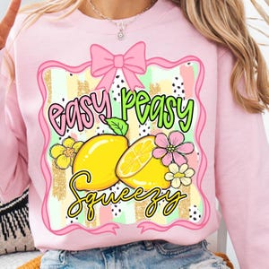 Może przedstawiać: Różowa bluza z nadrukiem przedstawiającym cytryny, kwiaty i napis "Easy Peasy Squeezy". Projekt zawiera różową kokardę i pasiasty wzór z złotymi brokatowymi akcentami.