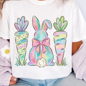 May include: White t-shirt featuring a pastel-colored bunny with a pink bow and two carrots. The bunny and carrots have a watercolor effect with gold accents. The design is perfect for Easter.