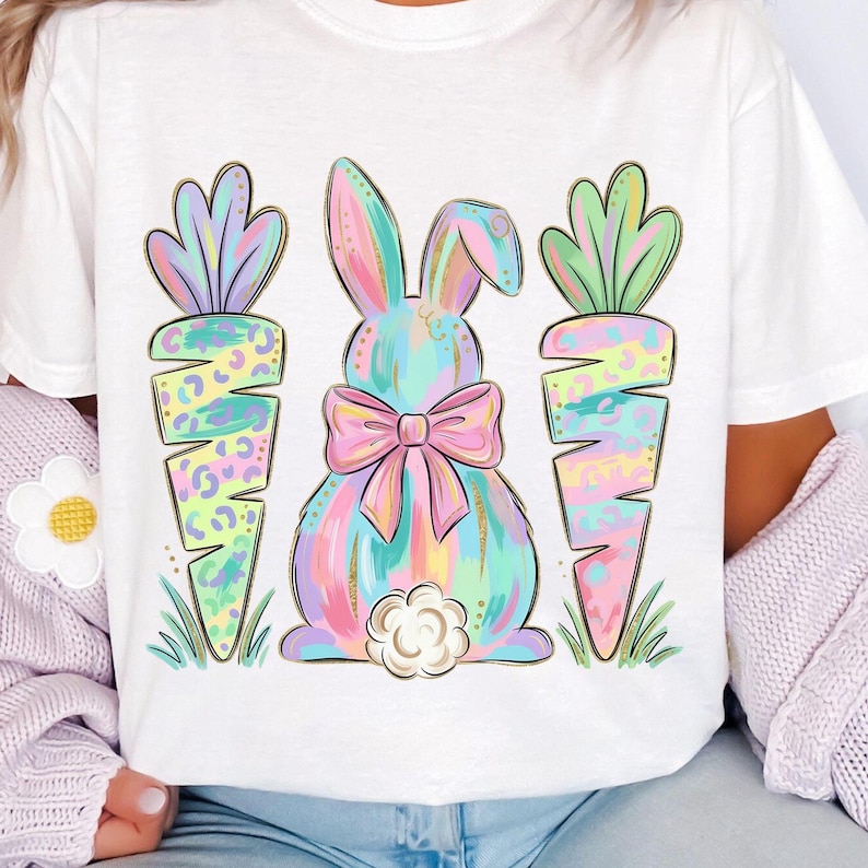 May include: White t-shirt featuring a pastel-colored bunny with a pink bow and two carrots. The bunny and carrots have a watercolor effect with gold accents. The design is perfect for Easter.