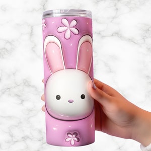 May include: A pink tumbler with a cartoon bunny design. The tumbler features a white bunny face with pink ears and a pink background with flower accents. The tumbler is held by a person's hand.