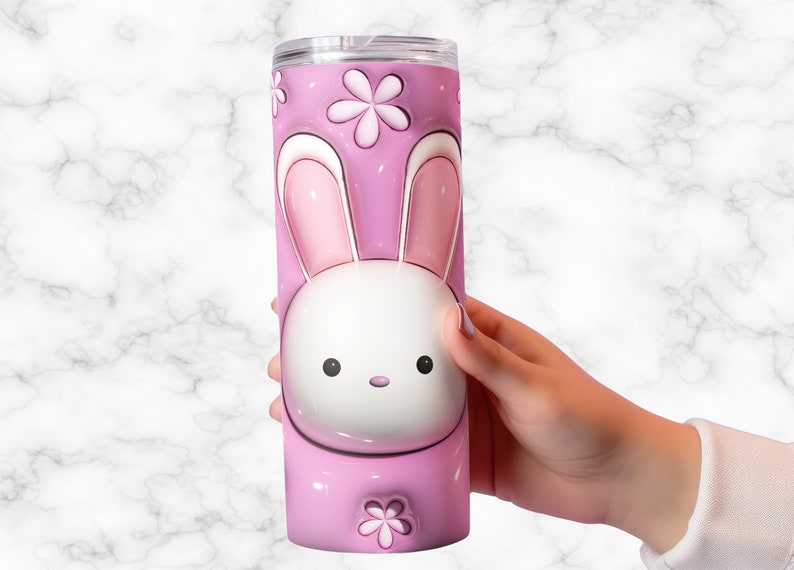 May include: A pink tumbler with a cartoon bunny design. The tumbler features a white bunny face with pink ears and a pink background with flower accents. The tumbler is held by a person's hand.