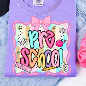 May include: A lilac-colored t-shirt with a colorful "Pre School" graphic. The design includes a pink bow, stars, daisies, an apple, a ruler, and a pencil. The shirt is size XL-TG-EG.