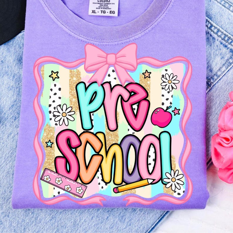 May include: A lilac-colored t-shirt with a colorful "Pre School" graphic. The design includes a pink bow, stars, daisies, an apple, a ruler, and a pencil. The shirt is size XL-TG-EG.