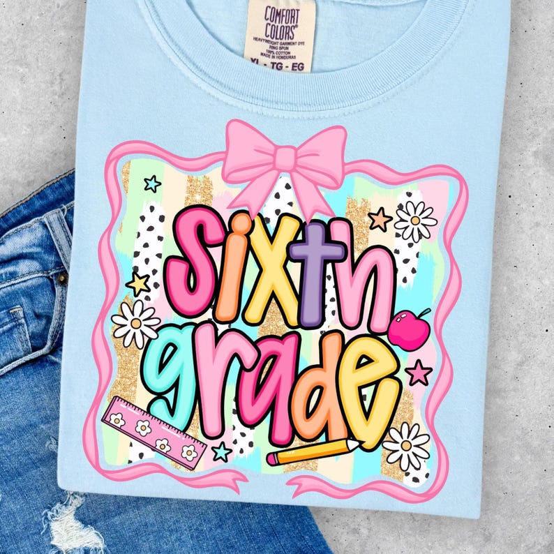 May include: Light blue Comfort Colors t-shirt with a colorful "Sixth Grade" graphic. The design includes a pink bow, stars, daisies, an apple, a ruler, and a pencil. The shirt is displayed on a pair of blue jeans.