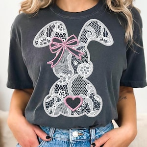 May include: A dark gray t-shirt with a white lace bunny design. The bunny has a pink bow and a pink heart on its chest.