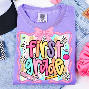 May include: A lilac Comfort Colors t-shirt with a colorful graphic that says "first grade." The design includes a pink bow, daisies, stars, a ruler, a pencil, and an apple. The shirt is laid on top of a pair of blue jeans.