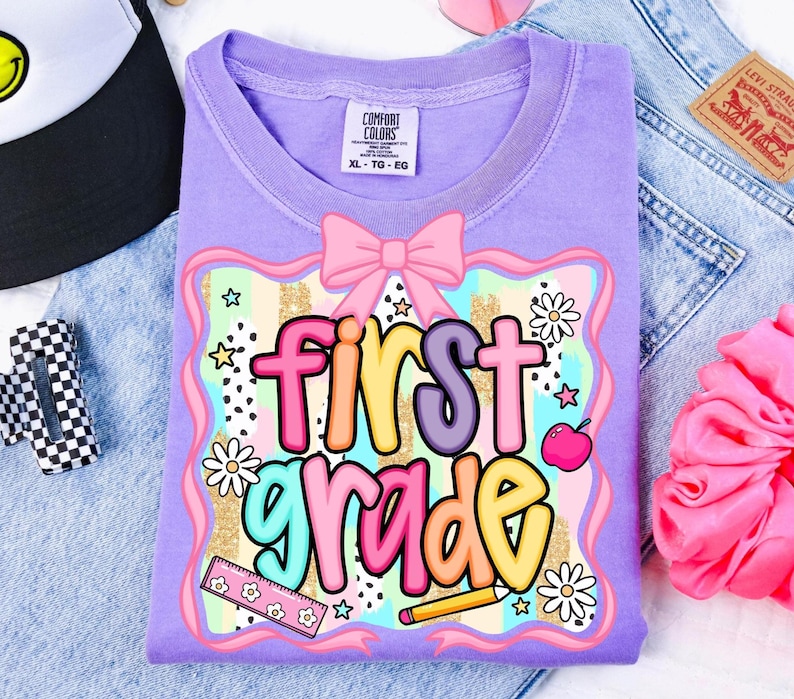 May include: A lilac Comfort Colors t-shirt with a colorful graphic that says "first grade." The design includes a pink bow, daisies, stars, a ruler, a pencil, and an apple. The shirt is laid on top of a pair of blue jeans.