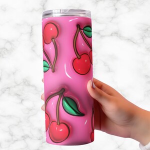 May include: A pink tumbler with a repeating pattern of red cherries with green leaves. The cherries appear to be 3D, with a glossy, raised effect. The tumbler is cylindrical and has a silver lid.